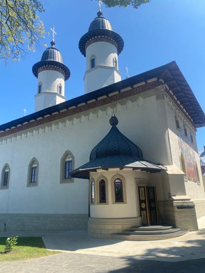 The image shows the exterior of a white church with two prominent dark, domed towers topped with crosses. The church has a tiled roof with ornate trim. There is a smaller, dark, domed entrance structure with arched windows and a decorative golden door. The facade features several arched windows with dark frames and a colorful mural on the right side. The building is situated in an outdoor area with a stone pathway, a grassy area with a small bush, and a clear blue sky. The scene is set in the city of Filioara, Romania, though no people are visible in the photograph.