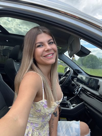 A young woman with blonde hair and a floral print dress sits in the driver's seat of a car. The car's interior is modern, with a large touchscreen display and a sunroof. The woman is smiling and looking at the camera. The exterior of the car appears to be in a rural or suburban setting, with green grass and trees visible in the background. It is daytime, and the sky is cloudy. The location is specified as Pastukhovo, Russia.