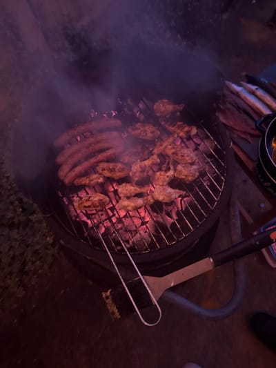 This image depicts an outdoor nighttime grilling scene at Neuville-aux-Bois, France, illuminated by the red glow of hot charcoal and ambient low light, casting a purplish hue. A black, round charcoal grill occupies the central and lower-left portion of the frame. On the grill rack, multiple items are cooking: approximately five to six long sausages are visible on the upper-left, while numerous pieces of chicken, primarily wings or drumettes, are spread across the right and lower sections of the grill. Visible smoke rises from the cooking meat and glowing red embers below the rack. A metal spatula with a black handle rests horizontally on the lower-right edge of the grill. A pair of metal tongs, one end integrated with a brush, is positioned across the lower-left section of the grill. To the far right, on what appears to be a dark wooden surface or table, several whole, slender fish are laid out. A dark cooking pot or pan is also partially visible on this surface, to the right of the fish. The ground surface around the grill is dark and indistinct. A dark shoe is partially visible at the bottom-right corner, indicating a human presence near the grill.