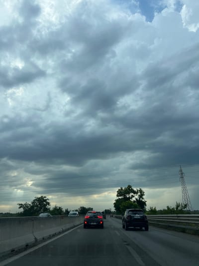 A vertical, eye-level, wide shot captures a view from a vehicle traveling on a highway, looking forward. The sky is filled with dramatic, dark gray clouds, suggesting an impending storm. The road ahead is asphalt with a white dividing line, and on the right, a metal guardrail separates the highway from a grassy embankment. A large, dark gray concrete barrier runs along the left side of the road. Several cars are visible on the highway, including a dark colored Fiat 500 in the foreground on the right lane, and a red car further ahead in the left lane. Beyond the guardrail, there are trees and vegetation. In the background, a tall, lattice-like electrical pylon stands prominently on the right. The overall scene conveys a sense of travel under brooding, overcast skies.