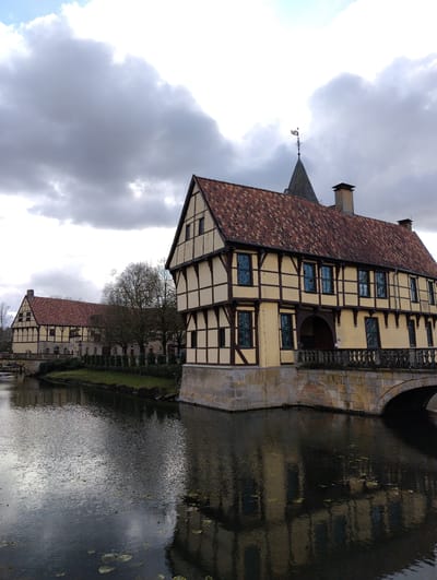 The image depicts a tranquil scene at a moated architectural complex in Burgsteinfurt, Germany, likely Schloss Burgsteinfurt. The primary focus is a large, two-story half-timbered building situated directly on a dark, rippled body of water, which reflects its facade and the overcast sky. This building features light cream-colored infill panels contrasted with dark brown timber framing, a prominent gabled red-tiled roof, and multiple windows with dark frames, many appearing tinted or reflecting blue hues. An arched stone bridge with an ornate balustrade connects the building's ground level to the adjacent land. Behind this structure, a dark grey conical spire topped with a weathervane is visible against the cloudy sky, alongside a stone chimney. To the left and further in the background, a second, similar half-timbered building with a red-tiled roof is present, also bordering the water. A narrow strip of green grass and manicured hedges line the water's edge between the two buildings, backed by several deciduous trees with bare branches. The sky is largely overcast with significant grey cloud cover and some brighter, lighter cloud formations. No individuals or discernible actions are present in the frame. The water surface contains scattered circular lily pad-like vegetation.