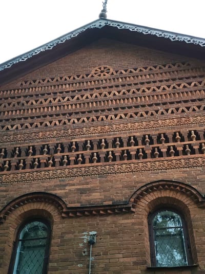 The image shows the brick facade of a building, likely a church or monastery, in Uglich, Russia. The facade is decorated with intricate patterns of brickwork, including geometric shapes and ornamental friezes. Two arched windows are visible at the bottom of the image, with reflections of trees visible in the glass. The sky is overcast.