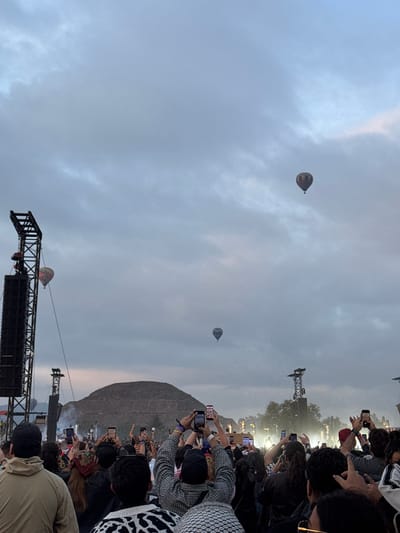 The image depicts a crowded outdoor event, likely a concert, with a large audience facing a stage. Many individuals are holding up their phones, presumably recording or taking pictures. In the sky, there are at least three hot air balloons visible. A large pyramid-like structure is noticeable in the background. Stage lighting and scaffolding are present. The general environment suggests an evening or dusk setting with a partly cloudy sky. The event could be taking place in San Francisco Mazapa, Mexico, given the presence of the pyramid-like structure which may indicate a location near ancient ruins.