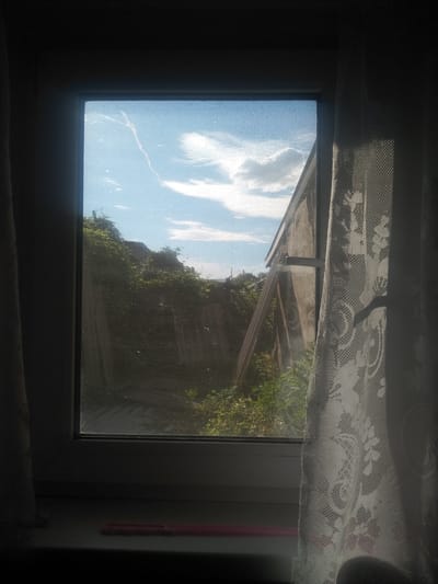 A view through a window shows a bright blue sky with wispy white clouds and a contrail. The window has a screen and a sheer white lace curtain on the right side. Outside the window, there is lush green foliage growing on a slope. A wooden structure, possibly a shed or fence, is visible to the right, partially obscured by the curtain. A narrow, pink object rests on the windowsill in the foreground, which is in shadow. The scene is lit by natural sunlight. The location is identified as Osakarovka, Kazakhstan.