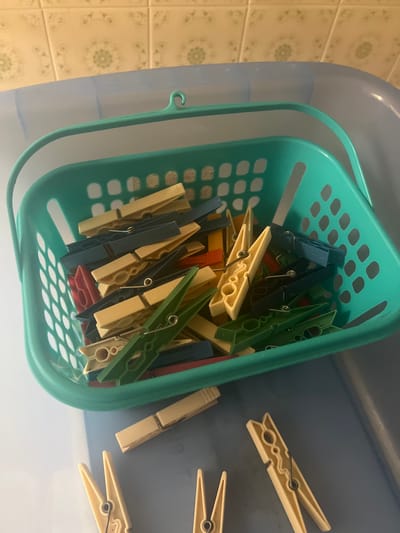 A collection of clothespins is organized in a teal-colored plastic basket. The basket is positioned within a larger, light blue plastic container. The clothespins are varied in color, including yellow, green, navy blue, and red. Some clothespins are also scattered on the surface of the light blue container. The background consists of tiled walls with a faint floral pattern. The scene appears to be indoors, possibly in a laundry room or storage area. There are no people visible in the image.
