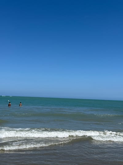 The scene depicts a beach in Cabarete, Dominican Republic. A vast expanse of clear blue sky occupies the left half of the image, contrasting with the ocean on the right. Two people are swimming in the ocean. Waves are visible close to the shore.