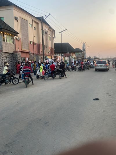 A street lined with buildings on the left is congested with motorcycles and people. A light-colored van is parked in the distance, further up the street. Utility poles extend alongside the buildings. The pavement appears gray and textured. The sky is a gradient of light colors, suggesting dusk or dawn. The scene conveys a bustling urban environment.