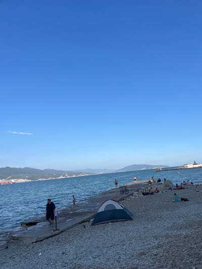 The image captures a scene at a rocky beach with the sea and a distant coastline under a clear blue sky. A man in a dark blue t-shirt and shorts stands on the paved area near the water, facing away from the camera. To his left, a small, partially set-up tent with red, white, and blue fabric is visible. A bicycle is leaning against the paved area near the tent.

Further along the paved walkway, several people are seen relaxing and sunbathing on the rocky beach. Some individuals are sitting or lying down, while others appear to be wading in the shallow water. The sea is calm with gentle ripples. In the background, a long pier or breakwater extends into the water, with structures at its end. The distant coastline features hills or mountains, and some buildings can be discerned along the shore.

The clear sky and bright lighting suggest it is daytime, likely late afternoon or early evening given the angle of the light. The presence of people in swimwear and engaging in beach activities indicates a warm, pleasant weather condition. The overall atmosphere appears relaxed and leisurely. No visible text is present in the image. The location is identified as Ivanovskaya, Russia.