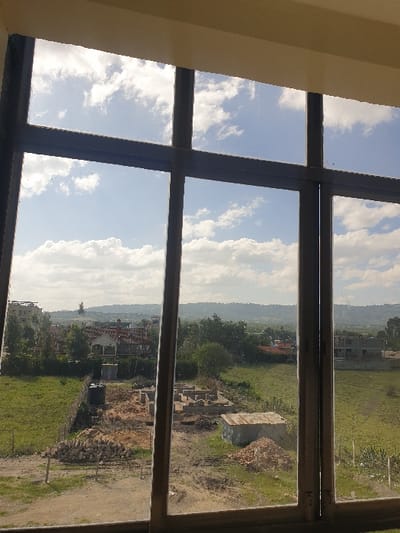 The image depicts an outdoor landscape viewed through a multi-pane window with dark grey mullions and transoms, exhibiting some surface dust. The upper portion of the view consists of a blue sky with numerous white cumulus clouds. The scene is situated in the city of Mumbuni North ward, Kenya. In the midground, a construction site is present, featuring a concrete block foundation, several distinct piles of earth and building materials (bricks/blocks), and a small, single-story structure with a corrugated metal roof. A black cylindrical water tank on a low stand is visible behind a wire fence in the left-midground. The surrounding environment comprises expansive green fields interspersed with scattered trees and bushes. The background reveals several residential buildings, including one multi-story structure under construction on the far right, with distant hills visible on the horizon. No individuals are present, and no discernible actions or interactions are occurring within the visible scene.