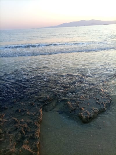 The image depicts a coastal scene at what appears to be sunrise or sunset. The foreground features rough, dark-colored rocks partially submerged in shallow, clear water. The rocks are irregular in shape and texture, with some areas reflecting light. Beyond the rocks, the water transitions to a calmer surface with gentle waves lapping at the shore. The middle ground shows the expanse of the sea, with subtle ripples and a line of foam from incoming waves. In the distance, a hazy mountain range is visible against a pale, softly colored sky, suggesting a peaceful, natural environment. The overall impression is one of tranquility. This scene is located in Saint Arsenius, Greece.