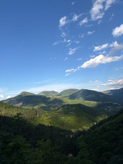 The image shows a landscape in Zasele, Bulgaria. There are green mountains with some houses nestled within the trees. The sky is blue with scattered white clouds. The sun is shining, casting shadows on the mountainside.