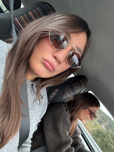 Two females are depicted inside a vehicle, likely in Le Cannet-des-Maures, France, as specified. The primary subject, positioned in the foreground and slightly to the left, has long brown hair and is wearing a light grey marled sweater. She wears large, reflective, gold-framed sunglasses with one lens partially obscured by the frame design, and her lips are slightly pursed. A dark grey seatbelt is visible across her chest. In the background, to the right and slightly lower, a second female is visible, also with brown hair, wearing a dark brown ribbed sweater and smaller, dark-lensed sunglasses. She is looking out the vehicle's window, which reveals green foliage and what appears to be a natural, possibly hilly or mountainous, landscape, consistent with a drive through a vegetated area. The car's interior includes a seat with a striped headrest featuring horizontal bands of brown, black, and muted colors. The scene suggests travel, with the primary subject taking a selfie and the secondary subject observing the external environment.