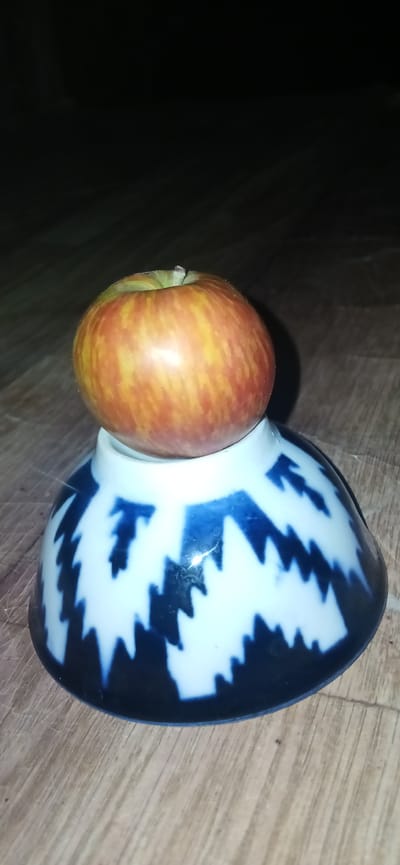 An apple with red and yellow streaks rests atop an inverted bowl with a blue and white zigzag pattern. The bowl is placed on a wooden surface. The background is dark. The scene is not identifiable as being in Kasansay, Uzbekistan.