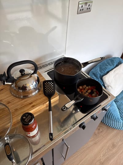 This overhead image captures a compact kitchen area with cooking in progress. Two black cooking pots are placed on a portable, two-ring electric hob. The pot on the left contains boiling water, while the pot on the right holds what appears to be meatballs and chopped red peppers being cooked.

Beside the hob, a shiny metal kettle sits on a wooden cutting board, next to a black slotted spatula. A glass jar of Loyd Grossman pasta sauce is also on the surface, with visible text including "GET FLAVOUR," "LOYD GROSSMAN," and "Check it!". Two glass pot lids are also present. The handles of the cooking pots bear the brand name "GO COOK."

The immediate surroundings include light wooden flooring to the right and a white wall to the left, featuring a British-style double wall socket. In the background, a blue ribbed furniture item, possibly a pet bed or small sofa, topped with a white pillow, is visible. The scene is well-lit, suggesting daytime. The wall socket confirms the location context of the United Kingdom.