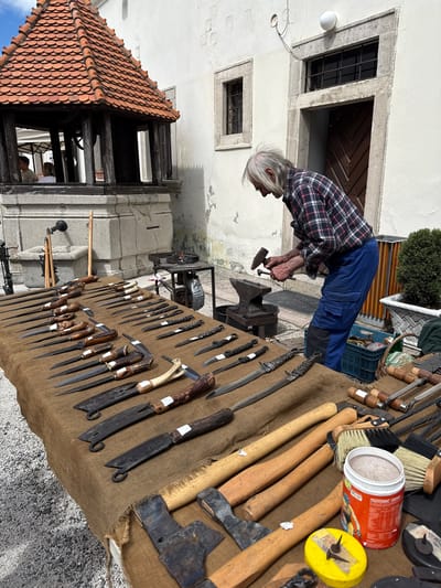 An outdoor scene captures an older craftsman, likely a bladesmith or blacksmith, actively engaged in his work at what appears to be a market or fair in Častá, Slovakia. The central figure is a man with gray hair, wearing a plaid shirt and blue work pants, bent over an anvil. He is holding a hammer, poised as if striking or shaping a piece of metal, demonstrating his craft.
In front of him, a large table covered with a brown, burlap-like fabric showcases a wide array of handcrafted bladed tools. These include numerous knives of various sizes and designs, some with intricate handles made from wood or antler, as well as several small axes or hatchets. The display suggests a sale or exhibition of his finished products. Among the items on the table are also brushes, possibly for cleaning or finishing, and a red container labeled "ISOTONIC DRINK" along with a yellow object that has small spikes or nails inserted into it.
The background features old, light-colored stone buildings with distinct stone window frames and a dark wooden door, characteristic of traditional European architecture. A tiled roof structure is visible on the left, further reinforcing the historical setting. Natural daylight illuminates the scene, suggesting a clear day. The man appears focused and absorbed in his work, embodying the dedication to his traditional craft. The overall atmosphere conveys a sense of a lively outdoor event centered around artisan skills.