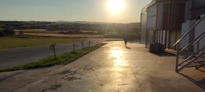 The image shows a person walking away from the camera on a concrete surface. The location appears to be in or near an industrial or agricultural area in Λάκκωμα, Greece. The sun is setting, casting a bright light and long shadows. The surrounding landscape includes fields, trees, a town, and distant hills. Part of a building with metal stairs and equipment is visible on the right. The scene suggests a late afternoon or early evening in a rural or semi-rural setting.