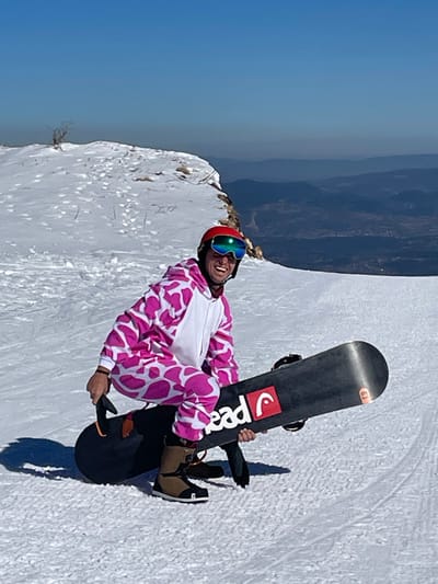 A male individual is depicted crouched on a snow-covered ski slope in Jahorina, Bosnia and Herzegovina, under a clear blue sky. He is wearing a pink and white cow-print full-body onesie, a red ski helmet, and reflective blue-green ski goggles. His mouth is open in a smile, showing teeth. He holds a black "Head" brand snowboard horizontally in front of him with both hands, balancing on brown ski boots. The immediate foreground consists of groomed snow. In the background, a snow-covered ridge with sparse vegetation is visible, leading to a panorama of hazy blue distant mountain ranges and valleys.