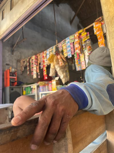 The image shows a hand holding onto a wooden structure, seemingly a part of a shop or small store in Paweden, Indonesia. The shelves inside display an array of snacks and food items. The hand belongs to an individual wearing a long-sleeved shirt with blue cuffs. Beyond the shelves, the floor area contains a mix of items, including a speaker and an orange container. The perspective suggests the image was taken from above, looking down into the shop.