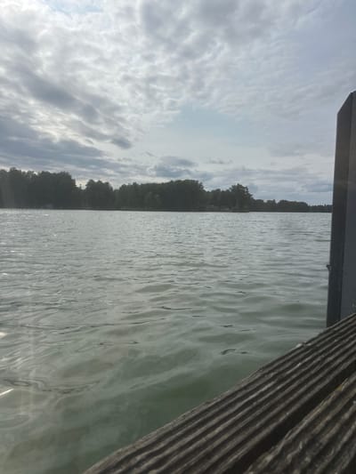 The image shows a view of a calm lake with a treeline on the opposite shore, under a cloudy sky. In the foreground, the edge of a wooden dock is visible. The image was taken from the dock, looking out over the water. The location is the town of Kloster Lehnin, Germany.