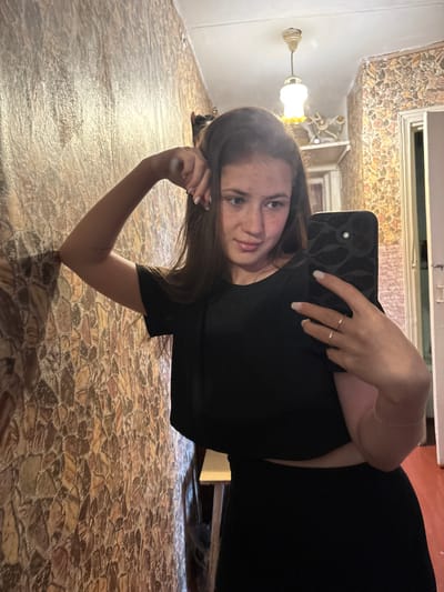 A young woman is taking a mirror selfie in what appears to be a residential hallway. She has long brown hair and is wearing a black crop top and black skirt. She is leaning against a textured wall with a pattern resembling stones. In the background, a light fixture hangs from the ceiling. The lighting suggests an indoor environment.