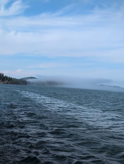 This is a wide shot of a body of water with a foggy coastline in the distance, under a cloudy blue sky. The foreground is dominated by dark, choppy water with small waves. The middle ground shows a dark, forested coastline. The far distance is obscured by a low-hanging fog that blankets the hills, with only the tops of some trees visible. A few small lights are visible in the distance along the coastline. The sky is a pale blue with streaks of white clouds. The scene appears to be captured from a moving vessel, as suggested by the wake in the water. The location is likely a coastal area in West Isles Parish, Canada, given the appearance of islands, fog, and evergreen forests typical of the region. There are no people or specific objects visible that would indicate a particular event or activity.