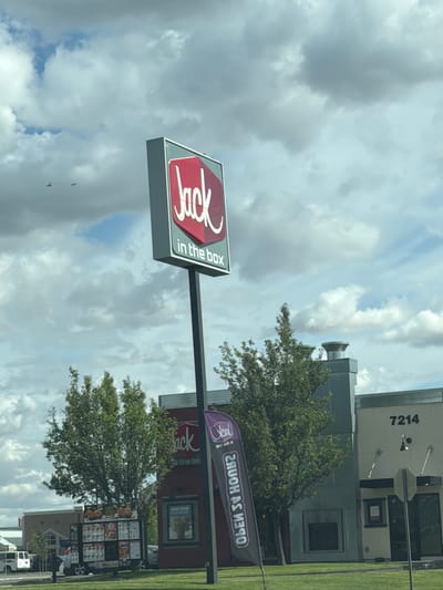 The image captures a Jack in the Box restaurant on a cloudy day in Pasco, Washington, United States of America. A prominent red and white Jack in the Box sign stands tall on a pole in front of the restaurant. To the left of the sign, a menu board displays various food items. To the right, a building with the number "7214" is visible, along with a dark awning and glass doors. A purple flag with the Jack in the Box logo is also present. The environment includes lush green trees and a paved area, likely a parking lot or drive-thru lane. The sky is filled with dramatic, scattered clouds.