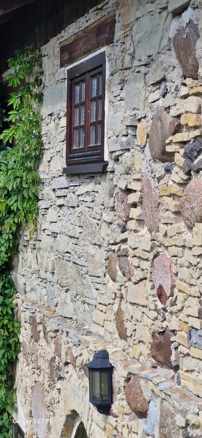 This is a close-up, eye-level, vertical shot of a stone wall of a building located in Pakruojis, Lithuania. The wall is constructed from irregular, rough-hewn stones of varying sizes and colors, predominantly light beige and grey, with some reddish-brown and darker grey stones interspersed. The stones are mortared together with grey cement.

To the left of the frame, a vine with lush green leaves is growing vertically up the wall. In the upper portion of the image, a wooden beam runs horizontally above a dark brown wooden window frame with multiple panes. The window is set into the stone wall.

Below the window, a black, traditional outdoor lantern is mounted on the wall. The lantern has a peaked top and glass panes. Part of an arched opening is visible at the bottom of the frame. The overall impression is one of rustic architecture. The image also contains a watermark in the bottom left corner that reads "Galaxy S23 FE".