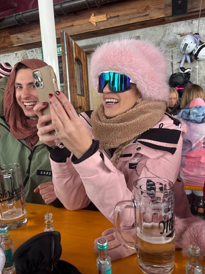 The scene depicts an interior setting, likely a bar or restaurant, in St. Christoph, Austria. Two individuals are seated at a rustic wooden table, engaging with the camera. The central figure is a female with fair skin, wearing a large, light pink faux fur hat, white-framed mirrored ski goggles with blue/green lenses, a brown faux-shearling neck gaiter, and a light pink ski jacket with black accents. She holds a light gold-colored smartphone horizontally with her left hand, which has red painted nails and a small tattoo, and is smiling broadly. To her left is a male with fair skin, wearing a brown head covering and a dark olive green jacket with "DOPE" visible. He is also smiling directly at the camera. On the wooden table, two clear glass beer mugs, partially filled with a light-colored beverage and foam, are visible. Several small clear glass bottles with silver caps and turquoise labels are also scattered on the table, alongside a black glove. In the background, a wooden ceiling beam with an arrow sign pointing left is present, along with a rustic wooden door, a white column, and a textured wall with ski helmets hanging on hooks. Other blurred individuals are partially visible in the background.