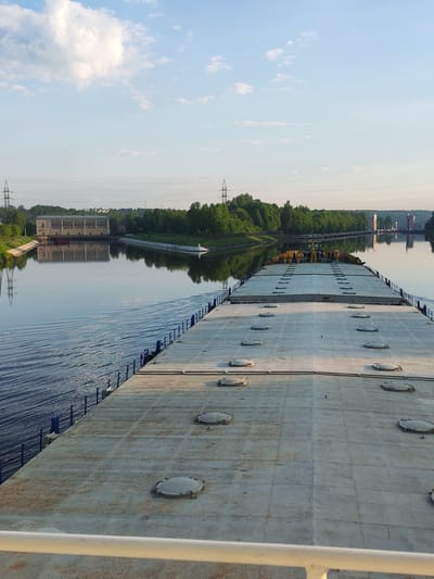 Here's a detailed description of the image:
The image appears to be taken from the deck of a large barge or ship traveling along a waterway near посёлок опытного хозяйства «Ермолино», Russia. The deck is gray, composed of rectangular sections with multiple circular access hatches. Blue railings run along the edges of the barge.
The waterway is wide, reflecting the sky. On the left bank, there's a large building, possibly a pumping station or industrial facility. A line of power transmission towers and lines run parallel to the waterway. Trees line both banks. A lock system is visible in the distance on the right.
The sky is blue with scattered white clouds, indicating a clear day. The lighting suggests it is either early morning or late afternoon.