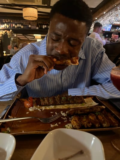 An adult male with dark skin, wearing a light blue striped long-sleeved shirt, is captured mid-action, consuming food in a restaurant. His eyes are closed, and his mouth is engaged with a piece of bone-in meat held in his left hand. The setting is identified as Misya Meze & Grill in Stevenage, United Kingdom.
On the wooden table in front of him, a rectangular brown ceramic plate displays his meal: a long skewer of ground meat (kebab style) resting on flatbread, accompanied by scattered white rice and a grilled half-tomato. Several pieces of grilled, bone-in chicken-like meat are also arranged on the plate. A silver fork rests amidst the rice. A glass containing a dark red liquid is partially visible on the right. In the immediate foreground, a small white square bowl contains a single, clean bone fragment.
The background reveals the interior of the restaurant, characterized by dark wooden ceiling beams and a cylindrical, patterned light fixture providing warm, ambient illumination. Shelves stocked with numerous wine bottles are visible on the left wall. Other blurred patrons are present, including a male in a white shirt near a bar area where glasses are suspended. The overall environment suggests an evening dining experience.