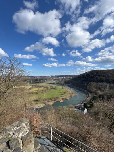 The image depicts a panoramic landscape view over the Ruhr River valley in Hattingen, Germany, from an elevated vantage point. The upper portion features a bright blue sky sparsely populated with white cumulus clouds. In the immediate foreground, to the bottom left, is a partial stone wall fragment, likely part of an observation structure, alongside a metal grated platform and railings descending out of view. Bare, leafless deciduous tree branches frame the left and right sides of the midground.

The midground is dominated by a serpentine river, likely the Ruhr, flowing from left to right. On the inner bend of the river, a large, irregularly shaped green field is visible, containing several scattered leafless trees and at least two prominent electricity pylons. Adjacent to the river's outer bend, a railway line runs parallel to the waterway. A distinct white two-story building with a dark roof stands near the river's edge on the forested slope to the right.

The background presents a distant urban or suburban area, identified as Hattingen, characterized by various buildings and infrastructure. A distinct area resembling a cemetery or industrial complex with numerous uniform structures is visible near the river in the mid-distance. The valley slopes and surrounding hills are covered with dense, bare deciduous forestation, with additional power lines visible across the landscape. No individuals or active subjects are discernible within the frame.