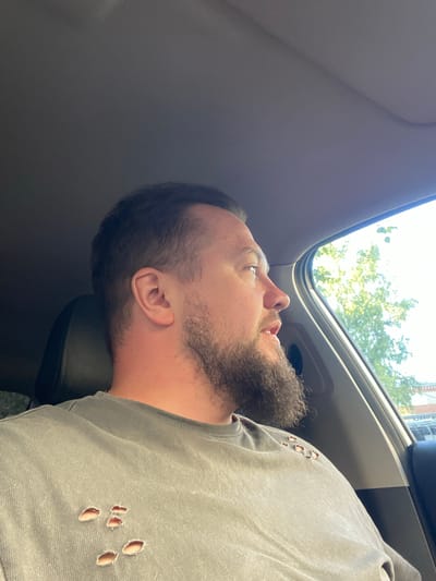 The image captures an adult male in a right-facing profile, seated within a vehicle in Sobinka, Russia. He is illuminated by bright natural light entering from the right side. The man has short, dark hair with a visible receding hairline and a full, dark brown beard. His mouth is slightly ajar, as if speaking or reacting. He is wearing a solid grey, short-sleeved crew-neck t-shirt featuring several irregularly shaped, distressed holes across the upper left chest and shoulder area. A dark grey seat headrest is visible behind his head, and the car's interior ceiling is a uniform dark grey fabric. Through the vehicle's right-side window, a bright outdoor scene under clear, sunlit conditions is visible. Lush green tree foliage dominates the immediate external view, suggesting a natural element within an urban or suburban setting. In the background, beyond the trees, elements of a building and an indistinct sign with faint lettering can be discerned, partially obscured by glare and foliage. No other individuals or interactions are visible.