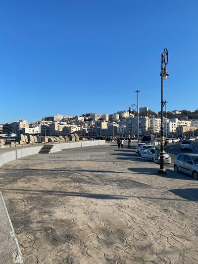 The image presents a view of the city of Beau Fraisier, Algeria from what appears to be a promenade or walkway. A clear, blue sky is visible above. The urban landscape is densely packed with buildings, primarily white in color, stacked along a hillside. Along the promenade, there are street lamps with distinctive circular features. Cars are parked along the side of the road next to the promenade. A few individuals can be discerned along the walkway. The walkway itself has a patterned, textured surface. A low concrete barrier is visible along the edge of the walkway.