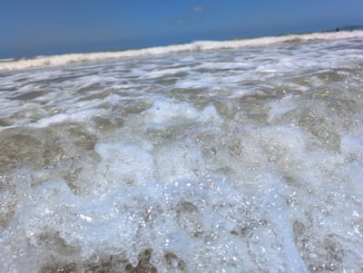 A close-up, low-angle shot captures the foamy crest of a breaking ocean wave. The water is clear and bubbly, with sunlight glinting off the surface, creating a sparkling effect. In the background, a distinct white line indicates the main body of the wave breaking, with a clear blue sky above. The horizon is slightly visible, and in the far distance, blurred figures of people can be seen on the shore. The scene is likely a beach in Tianya District, China, given the context. The focus is on the dynamic movement and texture of the water.