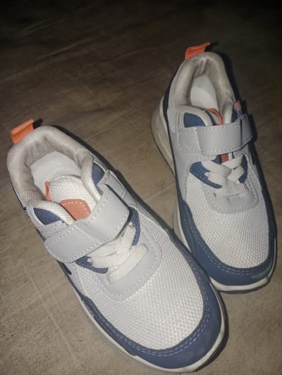 A pair of white and blue athletic shoes with orange accents is photographed from a high angle, lying on a light brown, textured surface. The shoes are facing upwards, with the left shoe positioned slightly behind and to the left of the right shoe. Both shoes feature white laces and a velcro strap across the midfoot. The right shoe is more in focus and shows a white mesh upper with blue accents around the toe and sides, as well as an orange pull tab at the heel. The background appears to be a wooden floor. The image is lit with a warm, dim light, casting some shadows. There is no indication of the location being in Meunasahara, Indonesia.