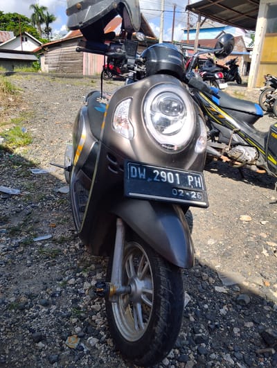 The image shows a front view of a grey scooter parked on a gravel surface in front of several wooden and metal-roofed buildings under a cloudy sky. The scooter has a black license plate with white text that reads "DW 2901 PH" and "02-26". A black helmet with "HONDA" written on it is placed on the handlebars. Behind the scooter, another motorcycle, possibly a Yamaha, is partially visible. The environment appears to be a semi-rural or informal urban area in Pompanua, Indonesia, indicated by the style of the buildings and the vegetation. There are no people visible in the image. The ground is covered in small stones and some scattered debris. The lighting suggests it is daytime.