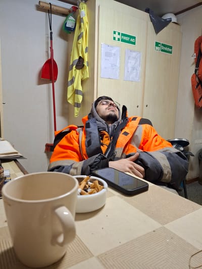 A man wearing glasses, a grey hooded sweatshirt, and an orange safety vest is sitting at a checkered table looking at a smartphone. Next to him is a mug and an ashtray filled with cigarette butts. Behind him is a wall with a first aid kit sign, a stretcher sign, and two pieces of paper attached to the wall. Below these items is a yellow reflective vest, a bottle of cleaner, and a red shovel. There are life vests on a shelf near the top of the frame. The location is possibly in Brunsbüttel, Germany.