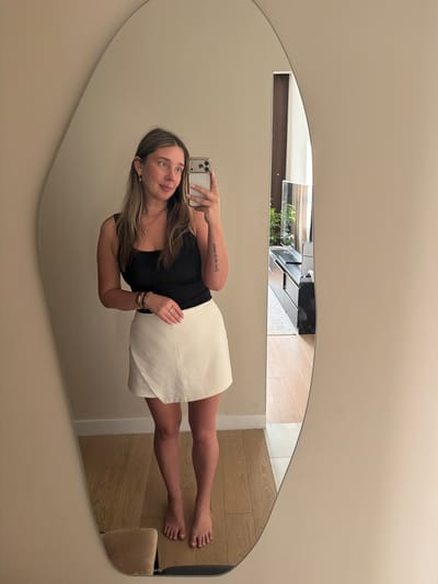 A woman stands in front of a large, irregularly shaped mirror, taking a selfie with her phone. She is wearing a black tank top and a white wrap skirt. She has blonde hair and is wearing earrings and bracelets. In the reflection of the mirror, a living room is visible, with a television, a black media console, and a large window. The floor is made of light-colored wood. The scene is indoors, likely in a residential setting. The specific location in Укромновское сельское поселение, Ukraine cannot be determined from the visual information provided.
