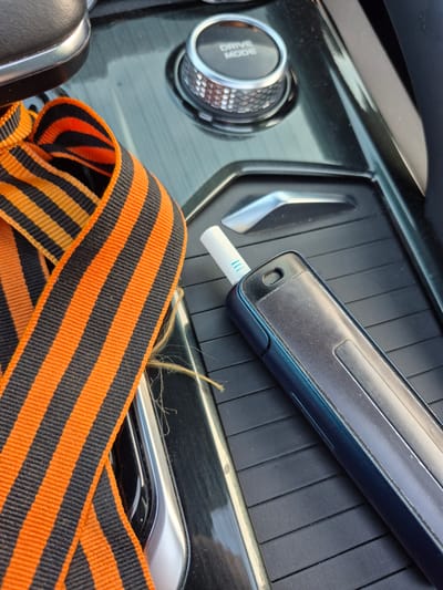 The image shows a close-up view of a car's center console. A dark blue electronic cigarette device with a white mouthpiece is resting on a textured black surface. To the left, a St. George's ribbon, characterized by its black and orange stripes, is draped across the console. In the background, a silver dial with the inscription "DRIVE MODE" is visible. The scene appears to be indoors, likely within a vehicle parked in Zapadny, Russia. There are no people present in the image.