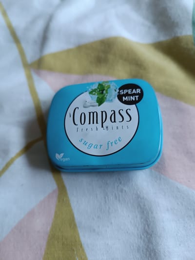 A close-up, overhead shot shows a small, rectangular, light blue tin of Compass brand spearmint, sugar-free mints. The tin is positioned diagonally on a fabric surface with a repeating geometric pattern of light yellow, pink, and white triangles. The lid of the tin features a circular white label with black text. The brand name "Compass" is written in large, stylized black letters across the center of the circle. Above "Compass," a small illustration depicts sprigs of mint leaves and crushed ice. Below "Compass," the words "Fresh Mints" are written in smaller black text. Further down, "sugar free" is written in a lighter shade of blue script. On the bottom left of the label, a small green vegan symbol is present. The background fabric is textured and appears to be a light blue-grey with faint white lines. No people are visible in the image. There is no information about the location of Ambert, France.