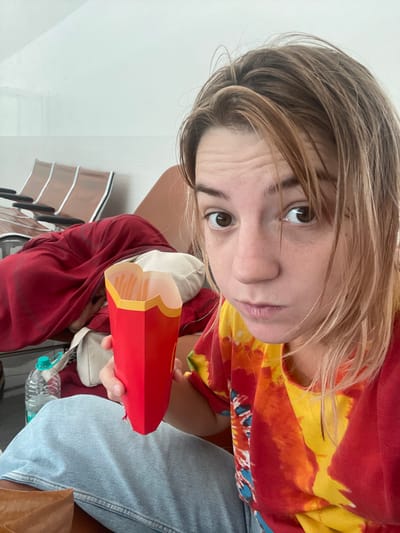 A young adult female is depicted in a forward-facing selfie-style photograph taken within a public waiting area, specifically identified as Muscat International Airport, Seeb Province, Oman. The subject has light brown, medium-length hair and brown eyes, looking directly at the camera with a neutral expression. She is wearing a red and yellow tie-dye t-shirt and light blue jeans. Her left hand holds a red McDonald's branded fries container, which is largely full of french fries. In the background, to the left, a row of airport-style waiting chairs with brown seats and black frames is visible. An adjacent seat is partially obscured by a draped red garment and a light-colored pillow. A clear plastic water bottle with a light blue cap rests on the floor next to the subject's left leg. The immediate environment features light-colored walls and the characteristic furnishings of an airport terminal.