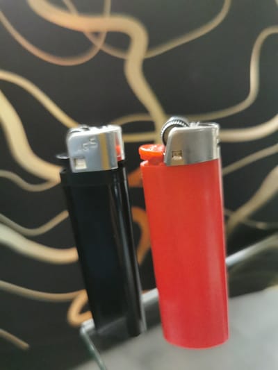 The image shows two lighters against a blurred background with a pattern of gold or yellow wavy lines on a dark surface. The lighter on the left is black with a silver top, and the text "AGS" is visible on the silver part. The lighter on the right is red with a silver top and a red igniter button. Both lighters are upright and positioned close to each other. The environment appears to be indoors, possibly on a reflective surface. There is no indication of a location in Bourg-lès-Valence, France, nor are there any people or actions depicted.