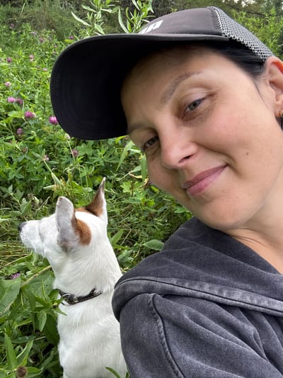 A selfie shows a woman wearing a black baseball cap and a dark gray hooded sweatshirt. She is smiling and looking at the camera. To her left, a white Jack Russell terrier with brown markings on its ears and head is looking to the left. Both the woman and the dog are outdoors, surrounded by green foliage and small pink flowers, suggesting a park or natural area. The setting is a daytime outdoor scene. The location is identified as Lipovaya I, Russia.