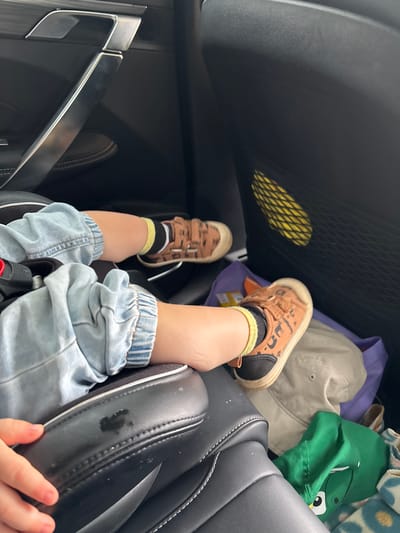 The image shows the lower legs and feet of a child sitting in a car seat. The child is wearing light blue denim jeans and brown sneakers with a cartoon pattern. The child's socks are yellow with black stripes. In the background, there is a black car door with a silver handle, and a mesh pocket containing a yellow object. There are also some items on the floor of the car, including a light-colored hat, a purple bag, and a green hat with a cartoon character. The image does not provide any information about the location in the city of Средняя Тойма, Russia.