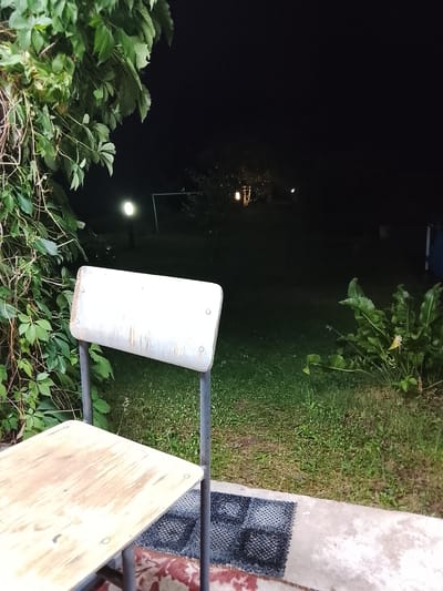 The image depicts a nighttime scene outdoors in Жалпақсай, Kazakhstan. In the foreground, a light-colored wooden table and chair are positioned on a concrete surface. The chair has a metal frame and a worn, white seat. A woven rug is placed on the ground between the table and chair.
Behind the furniture, a grassy area stretches into the background, illuminated by a distant streetlight. Further back, the faint glow of lights suggests structures or an event taking place in the distance. Lush green foliage from a climbing plant covers the left side of the frame, contrasting with the dark night sky. The overall impression is one of a quiet, possibly rural, evening setting. No people or text are visible in the image.