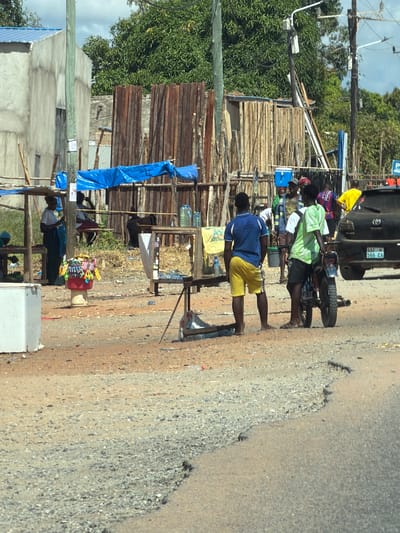 A roadside scene in Muxara, Mozambique shows a paved road with a rough, degraded edge. Along the edge of the road, several people are standing barefoot. One man in a blue shirt and yellow shorts is walking. Another man in a green shirt is standing next to a small motorcycle. Behind the motorcycle is a black SUV with a visible license plate number (AAF 506 CA). Adjacent to the road is a makeshift market with wooden structures and blue tarps. Various items are displayed for sale. There is vegetation and trees in the background. Utility poles are visible overhead. The overall scene appears to depict typical roadside activity in a developing area.