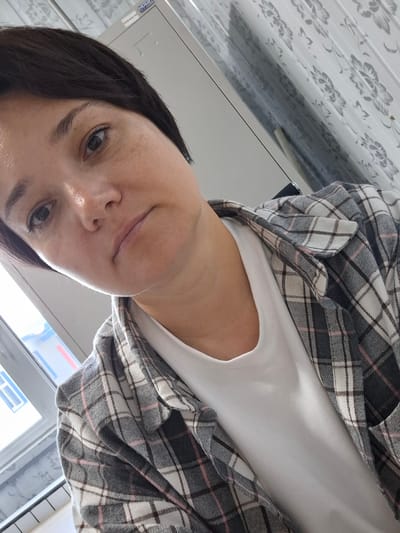 A woman with short dark hair and light skin with freckles is taking a selfie in what appears to be an office or industrial setting. She is wearing a white t-shirt under an open plaid shirt in shades of gray, white, and pink. The background includes a filing cabinet and a window with blinds. The room has a floral patterned wallpaper. The overall lighting suggests an indoor environment with natural light coming from the window. The location is identified as Tecnimont camp, Russia.