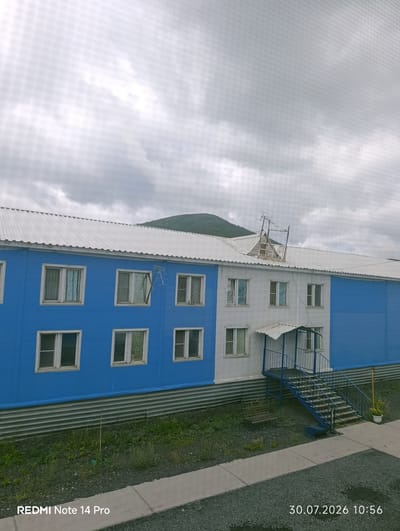The image shows a two-story building with a white, sloped roof, located in the settlement of Omchak, Russia. The building's exterior is painted with two distinct color sections: the lower half is a vibrant blue, and the upper half is white. Each story has a row of rectangular windows with white frames. A blue staircase with metal railings leads to a covered entrance on the white section of the building. An antenna structure is visible on the roof towards the center of the building. The foreground consists of a gravel path and some sparse vegetation. In the background, a gently sloping, tree-covered hill is visible under a cloudy sky. The timestamp "30.07.2026 10:56" and "REDMI Note 14 Pro" are present in the lower right and left corners, respectively. There are no people visible in the image.