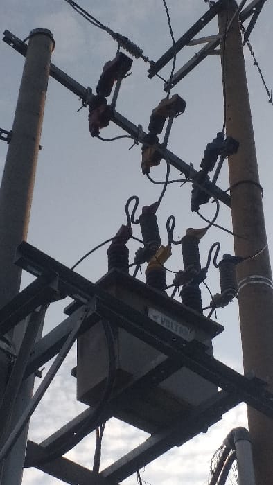 A low-angle, forensic view depicts an electrical power distribution setup in Lamreung, Indonesia. The scene is dominated by two utility poles supporting a suspended electrical transformer. The left pole appears concrete and dark grey, while the right pole is also a utility pole, slightly lighter in tone. A large, rectangular, dark grey electrical transformer, marked with "VOLTRON" on a label, is centrally mounted on a robust black metal framework positioned between the two poles. Overhead metal cross-arms extend from both poles, supporting multiple black, yellow, and brown ceramic or polymer insulators and associated high-voltage fuses or disconnect switches. Numerous black electrical wires connect these components, leading into and out of the transformer. The background consists of a predominantly overcast or cloudy sky, with some lighter patches indicating partial cloud cover. No individuals or active movements are observed.