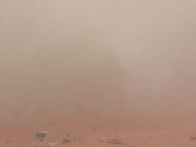 The image shows a hazy, dusty environment, likely during a dust storm, which is common in parts of Nigeria, including Nanka. The scene is dominated by a thick, reddish-brown haze that obscures visibility. In the foreground, the ground appears to be sandy or dusty, with some scattered debris, possibly including a piece of white plastic. The overall impression is one of low visibility and poor air quality due to the dust.